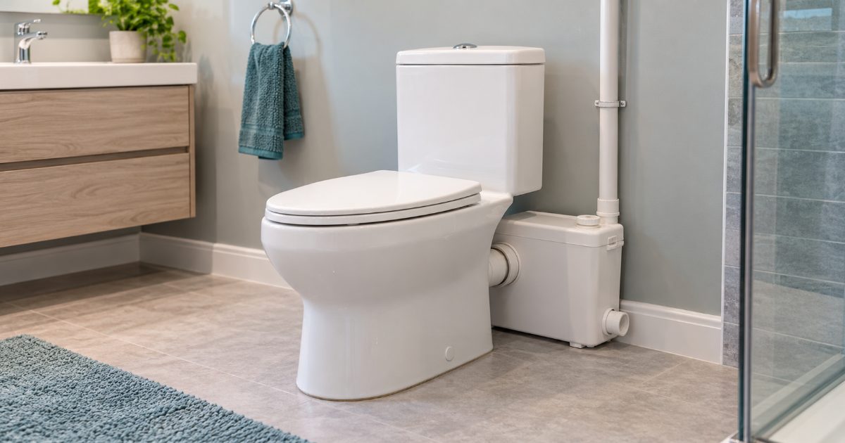 A white two-piece toilet in a basement bathroom with a compact white macerator box behind it and a narrow white discharge pipe running up the wall