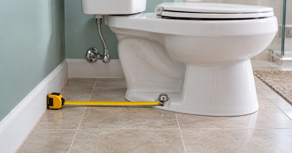 A yellow tape measure stretched along a tiled floor from the wall behind a white two-piece toilet to the chrome closet-bolt cap at its base